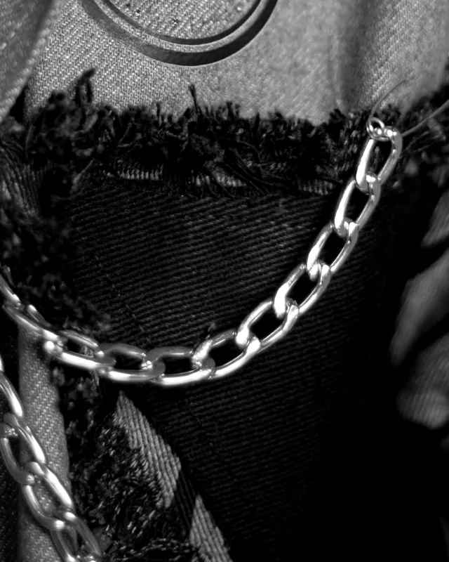 Close-up of frayed black denim set into a jacket front, with a silver chain looped across it.
