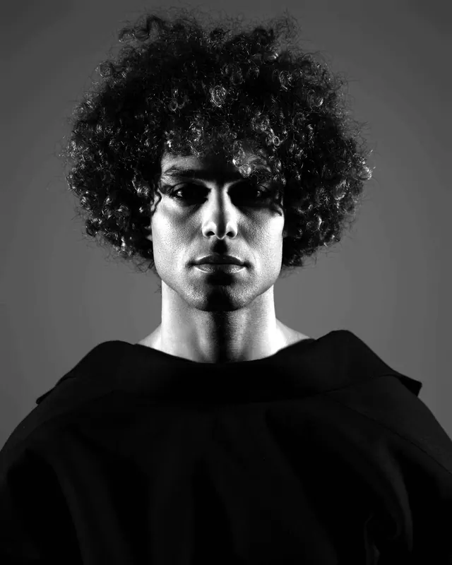 A man with curly hair in a heavy black hooded top, lit from the front.