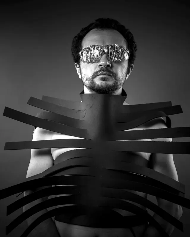 A man in reflective glasses wearing a harness of black strips that wrap his torso like ribs.