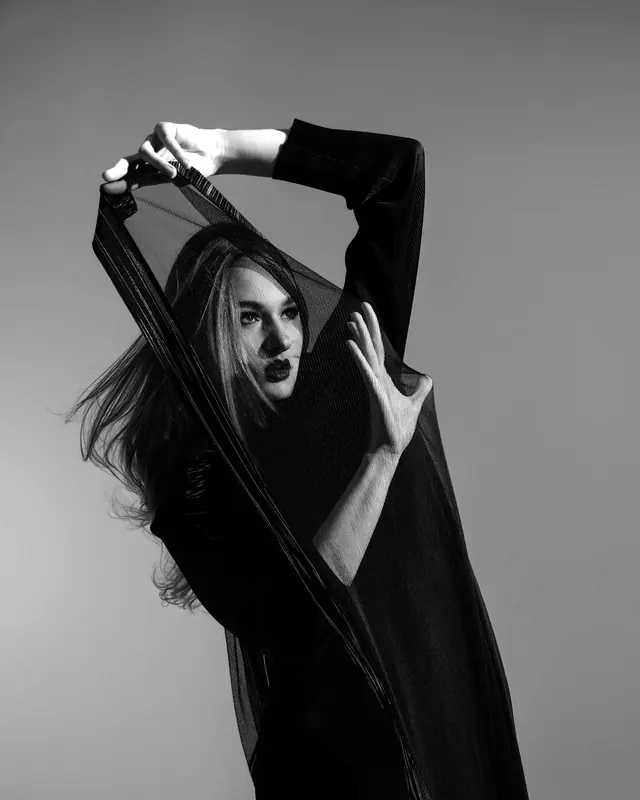 A model raising both arms inside a long black dress that falls in ribbons.